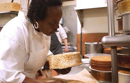 A baker carefully inspects a cake in a busy kitchen, surrounded by various baked goods and equipment.