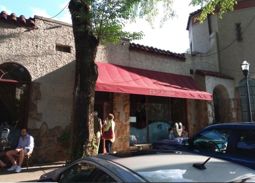 A restaurant with a red awning, "Chez Fontfont," surrounded by trees and parked cars, with people sitting outside.