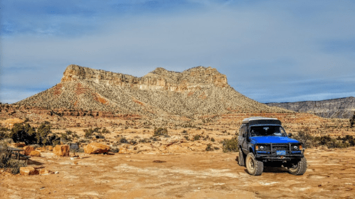 A blue SUV parked on rocky terrain with a large, rugged mountain in the background under a clear sky.