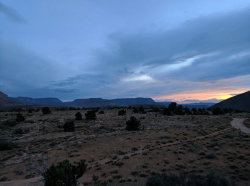 A serene landscape at dusk, featuring mountains, a cloudy sky, and a winding dirt path through sparse vegetation.