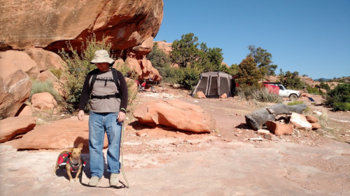 A person stands with a dog in a rocky outdoor area, with tents and vehicles in the background under a clear blue sky.