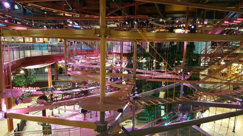 A colorful indoor ropes course with multiple levels and platforms, surrounded by vibrant lighting and greenery.