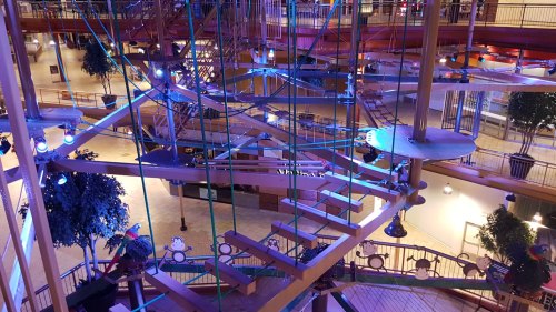A colorful indoor play structure with ropes and platforms, surrounded by trees and ambient lighting.
