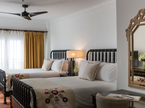 A cozy hotel room featuring two beds, a lamp, a mirror, and bright curtains, with floral decor and warm lighting.