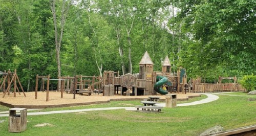 A wooden playground with slides, swings, and climbing structures, surrounded by trees and grassy areas.