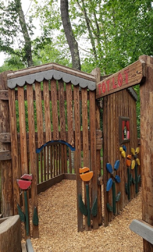 Wooden play structure with colorful flowers, labeled "TOT LOT," surrounded by trees and wood chips.