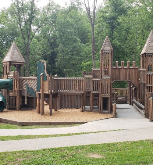 A wooden playground with towers, slides, and climbing structures, surrounded by trees and a grassy area.