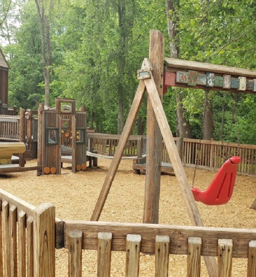 A wooden playground with swings and climbing structures, surrounded by trees and covered in wood chips.