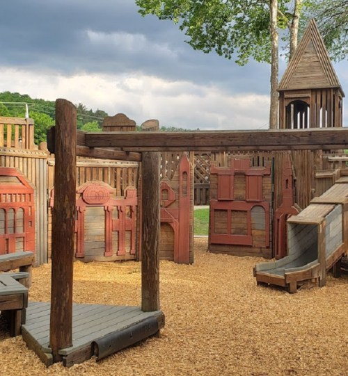 A wooden playground with slides and play structures, surrounded by trees and a cloudy sky.