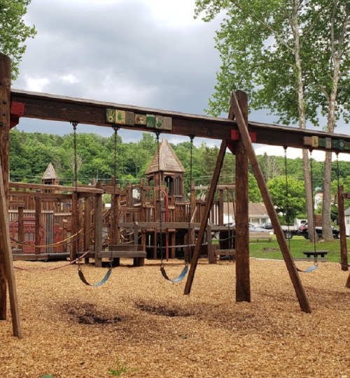 A playground with wooden swings and a large play structure surrounded by trees and cloudy skies.