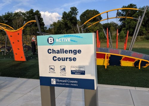 Sign for a challenge course at a park, stating rules and emergency contact information, with playground equipment in the background.