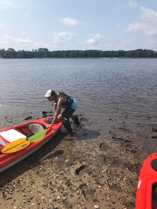 A person in a kayak prepares to launch into a calm river on a sunny day, with trees in the background.