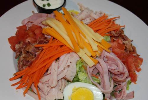 A colorful salad topped with sliced meats, cheese, carrots, tomatoes, bacon, and a hard-boiled egg, served with dressing.