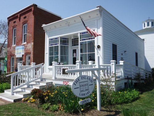 A charming white library building with an American flag, surrounded by greenery and flowers, inviting visitors inside.