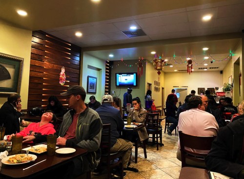 A busy restaurant scene with diners at tables, a TV on the wall, and festive decorations in a warm atmosphere.