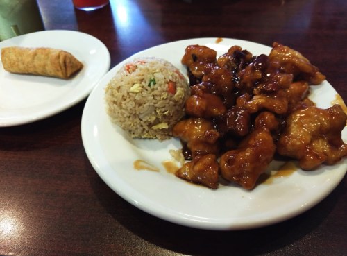 A plate of sweet and sour chicken with fried rice, accompanied by a spring roll on a separate plate.