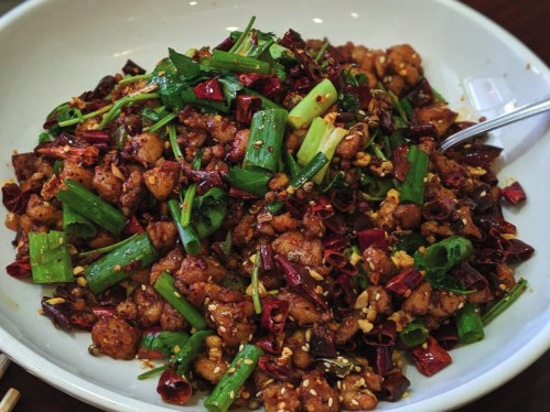 A bowl of spicy stir-fried chicken with green onions and dried red chilies, garnished with sesame seeds.