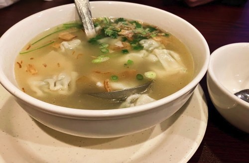 A bowl of clear soup with dumplings, green onions, and crispy toppings, served with a small spoon on the side.