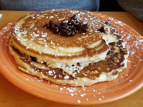 A stack of fluffy pancakes topped with chocolate chips and powdered sugar on an orange plate.