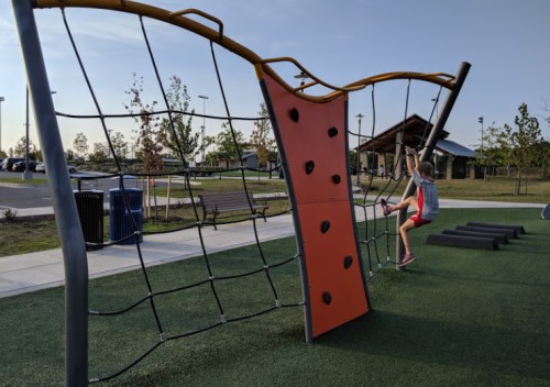 A child climbs a colorful play structure at a park, with benches and trees in the background under a clear sky.