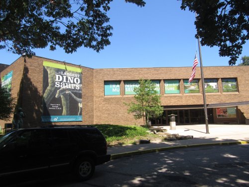 Exterior of a museum building featuring banners about dinosaurs and a flag, surrounded by trees and clear blue sky.