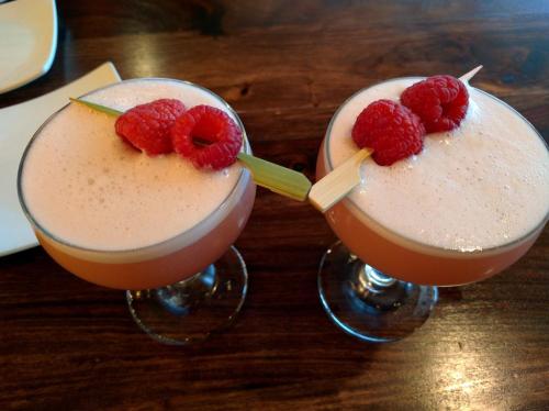 Two cocktails with frothy pink tops, garnished with raspberries and a piece of lemongrass, on a wooden table.