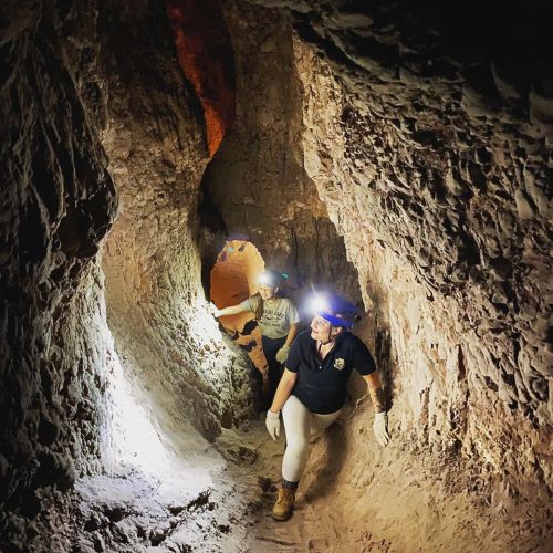 Two people explore a narrow cave passage, wearing headlamps and gloves, with rocky walls illuminated by their lights.