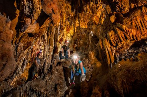 Three people explore a cave, wearing helmets with headlamps, surrounded by stunning rock formations and illuminated walls.