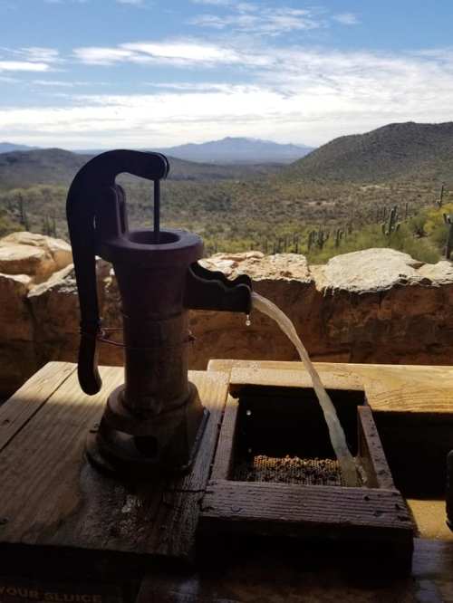 A vintage water pump with flowing water, set against a scenic desert landscape and distant mountains.