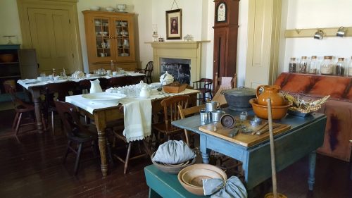A historic kitchen with wooden tables, chairs, and various cooking utensils, showcasing a rustic, vintage atmosphere.
