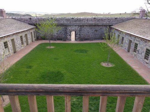 A grassy courtyard surrounded by stone buildings, with trees and a pathway leading to an archway in the background.