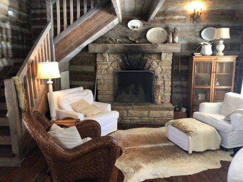 Cozy rustic living room with a stone fireplace, wooden stairs, and comfortable seating on a fur rug.