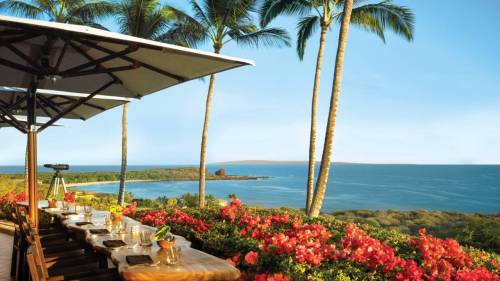 A scenic view of a coastal landscape with palm trees, vibrant flowers, and a dining table overlooking the ocean.