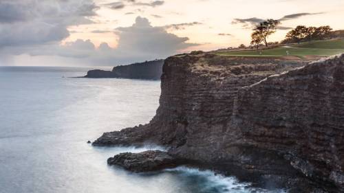 A scenic coastal view featuring rugged cliffs, gentle waves, and a sunset sky with scattered clouds.