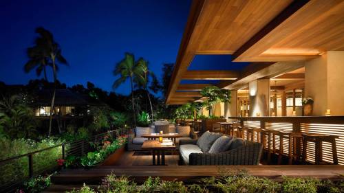 A stylish outdoor lounge area at night, featuring comfortable seating, plants, and soft lighting under a wooden canopy.