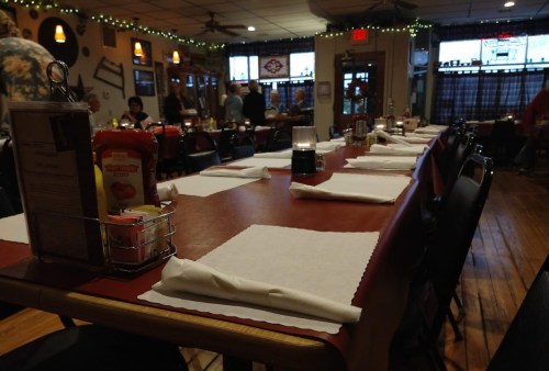 A long dining table set with napkins and condiments in a cozy restaurant, with patrons in the background.
