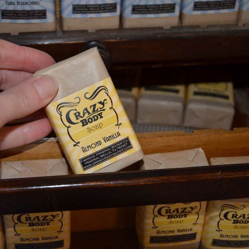 A hand holding a bar of Crazy Body Soap in Almond Vanilla, with more soap bars displayed in the background.