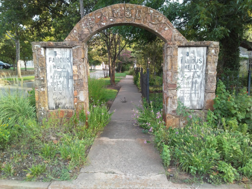 Stone archway with weathered signs reading "Famous Well" and "Keep Well with a Mission," surrounded by greenery and flowers.