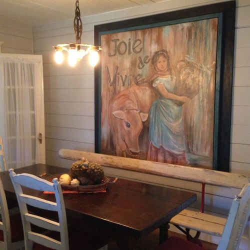 A cozy dining area featuring a wooden table, chairs, and a large painting of a girl with a cow. Warm lighting adds charm.