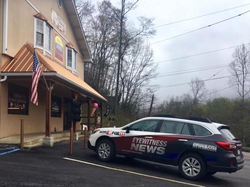 A news vehicle parked outside a building with an American flag, surrounded by trees and a cloudy sky.