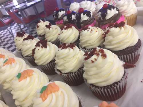 A variety of decorated cupcakes with swirled frosting, including red velvet and colorful toppings, displayed on a tray.