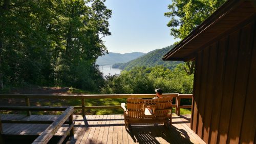 A person sits on a wooden bench overlooking a serene lake surrounded by green hills and trees.