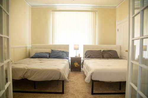 A cozy bedroom with two beds, light yellow walls, and a lamp on a nightstand between them. Natural light from a window.