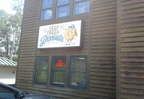 Sign for Deep Creek Donuts on a wooden building, featuring a cartoon donut and an "Open" and "Hot Donuts" neon sign.