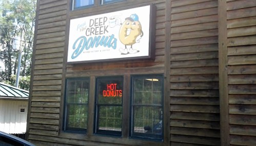 Sign for Deep Creek Donuts on a wooden building, featuring a cartoon donut and a "Hot Donuts" neon sign in the window.