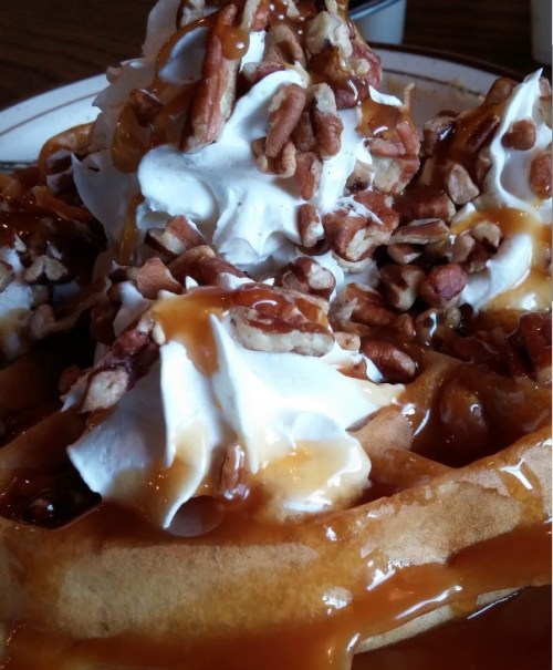 A close-up of waffles topped with whipped cream, caramel sauce, and chopped pecans.