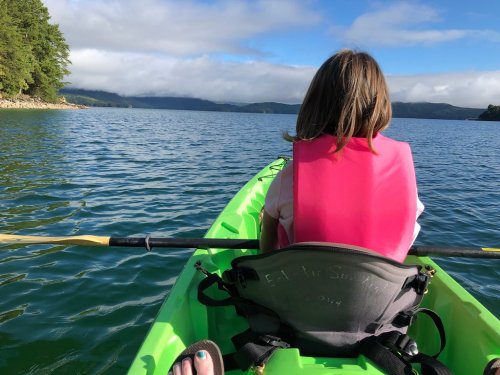 A person in a pink life jacket sits in a green kayak on calm water, surrounded by trees and mountains under a blue sky.