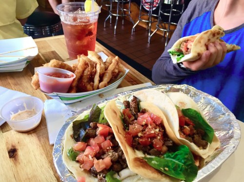A plate with three tacos filled with meat and toppings, alongside a basket of fried food and a drink.