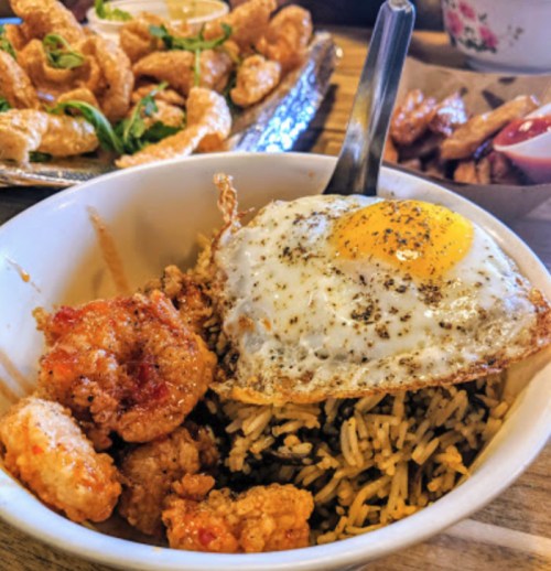 A bowl of rice topped with a fried egg and shrimp, with fried snacks in the background.