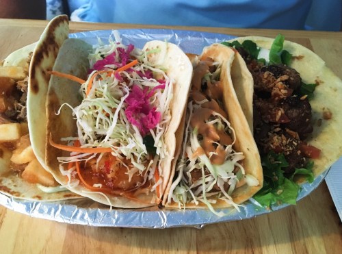 A platter of four tacos filled with various toppings, including cabbage, carrots, and sauces, served on a silver tray.
